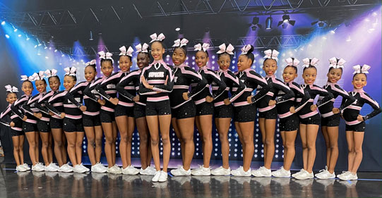 Nineteen girls in cheer uniform standing on a stage under purple and blue light. 
