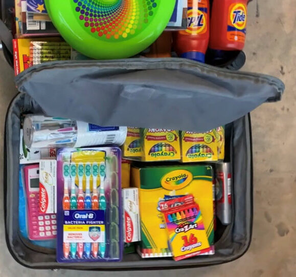 A suitcase full of toothbrushes, calculators, crayons, and toothpaste. Other various supplies including a bottle of Tide sit nearby. 